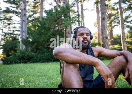 Ein afrikanischer Athlet mit Kopfhörern sitzt auf einem grasbewachsenen Feld. Er trägt ein schwarzes Tanktop und Shorts Stockfoto