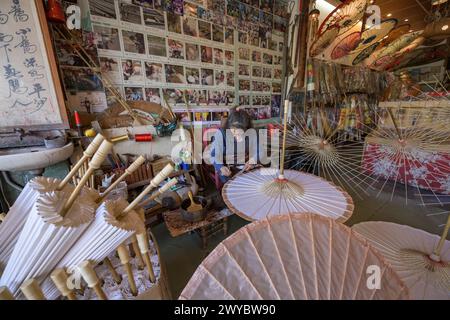 In einer Werkstatt konzentriert sich ein Handwerker auf die Herstellung traditioneller chinesischer Regenschirme, umgeben von fertigen Produkten Stockfoto