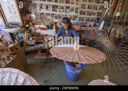 In einer Werkstatt konzentriert sich ein Handwerker auf die Herstellung traditioneller chinesischer Regenschirme, umgeben von fertigen Produkten Stockfoto