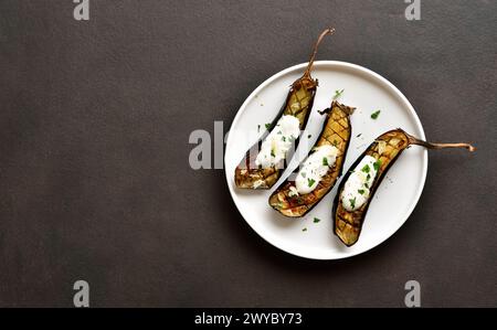 Gebackene Aubergine mit Knoblauch-Joghurt-Dressing auf Teller über dunklem Hintergrund mit Kopierraum. Draufsicht, flach Stockfoto