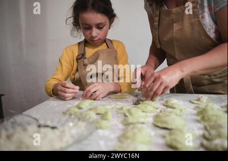 Hübsches kleines Mädchen in beigefarbener Kochschürze, das am bemehlten Küchentisch steht und ihrer Mutter hilft, Klöße zu stopfen und zu Formen. Mama und Tochter kochen Stockfoto