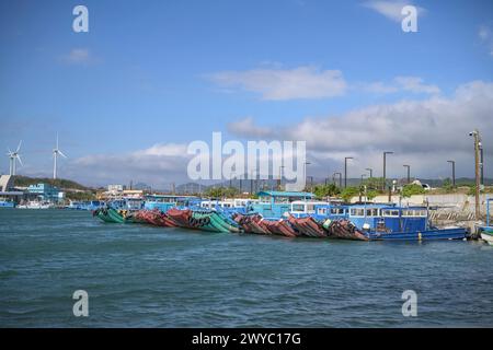 Blaue Fischerboote liegen in einem Hafen mit ruhigem Wasser und Windturbinen im Hintergrund Stockfoto