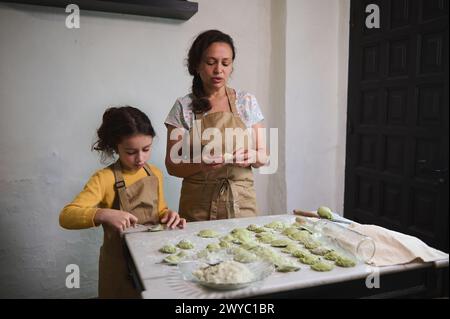 Hübsches kleines Mädchen in beigefarbener Kochschürze, das am bemehlten Küchentisch steht und ihrer Mutter hilft, Klöße zu stopfen und zu Formen. Mom und Dughter kochen Stockfoto