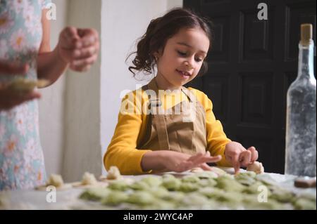 Entzückendes kleines Mädchen in beigefarbener Schürze, das mit ihrer Mutter Knödel zubereitete, am bemehlten Tisch in der rustikalen Küche des Landhauses stand. Familien-Tradi Stockfoto