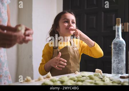 Weißes entzückendes, schelmiges kleines Mädchen in beigefarbener Schürze, lächelt beim Verkosten von Kartoffelpüree, während sie mit ihrer Mutter in der Ho Klöße kocht Stockfoto