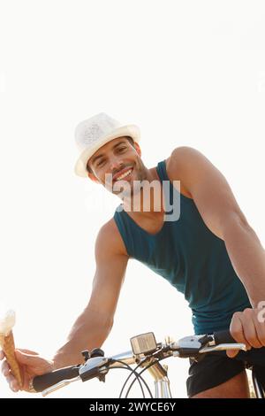 Mann-, Eis- und Fahrradtour für Reisen im Freien und Entspannung für eine Pause auf weißem Hintergrund. Junge Person, lächeln und sitzen auf dem Fahrrad mit Dessertkegel für Stockfoto