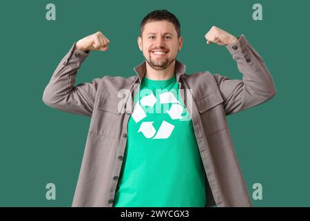 Glücklicher junger Mann im T-Shirt mit Recycling-Logo, das Muskeln auf grünem Hintergrund zeigt. Ökologisches Konzept Stockfoto