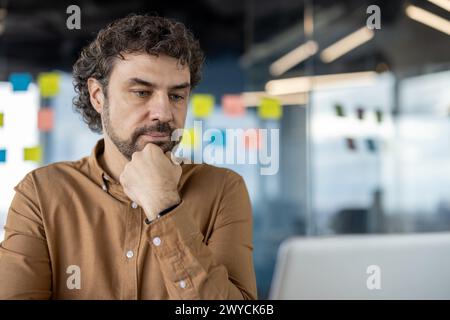 Nachdenklicher hispanischer Geschäftsmann, der tief fokussiert und besinnlich ist, während er in einem modernen Büroumfeld Hand am Kinn sitzt. Stockfoto
