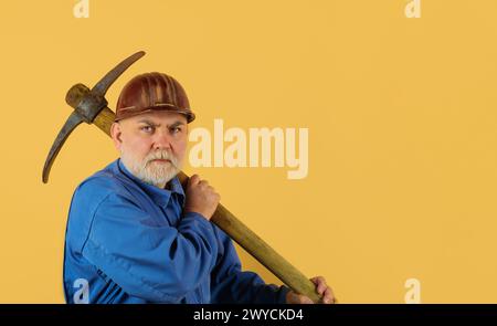 Ernsthafter bärtiger männlicher Baumeister in Overalls und Schutzhelm mit Meißelaxt. Bauwerkzeuge. Handwerker, Bauunternehmer oder Bergarbeiter in Uniform und Schutzhelm Stockfoto