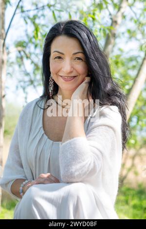 Lächelnde Frau mittleren Alters, mit langen dunklen Haaren, in weißer Bluse, in sonniger Natur. Verströmt eine ruhige Atmosphäre der Selbstfürsorge und gut b Stockfoto