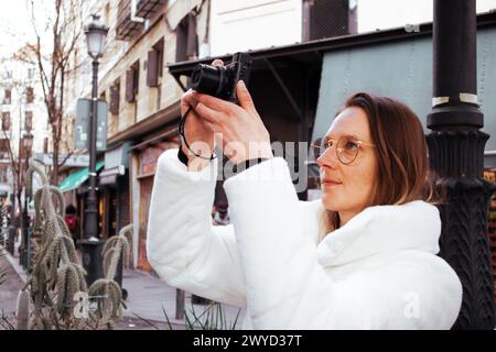 Madrid, Spanien. 1. März 2024. Eine junge Frau in einem weißen Pelzmantel macht ein Foto mit einer kleinen Digitalkamera auf einer Stadtstraße. Reisefotografin. Stockfoto