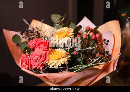 Lebhafter Blumenstrauß mit roten Rosen, gelben Chrysanthemen und Beerenzweigen, gewickelt in Pfirsichpapier für festliche Anlässe. Stockfoto
