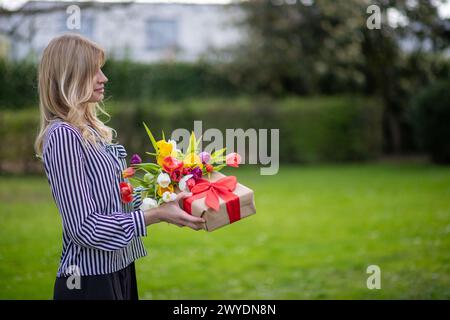 Eine junge Frau, die Blumen und Geschenke im Freien hält. Stockfoto