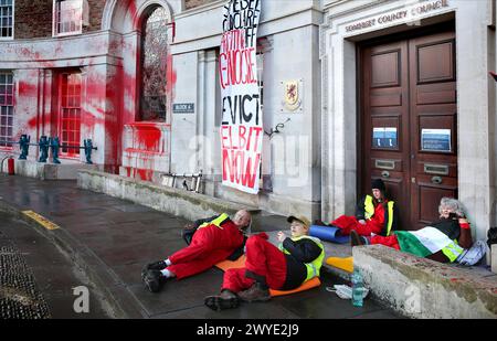 Palästinensische aktionäre sitzen in Zweiergruppen bei einer Demonstration vor dem Rathaus des Bezirks Somersetís. Unterstützer der Palästinensischen Aktion blockieren den Eingang zur County Hall mit Sperren und sprühen rote Farbe auf das Äußere des Gebäudes. Die Demonstranten fordern, dass der Stadtrat von Somerset den Mietvertrag für Büroflächen beendet, die sie an Elbit Systems in Aztec West, Bristol, vermieteten. Sie argumentieren, dass die von Elbit Systems in Großbritannien hergestellten Waffen von der israelischen Verteidigungsstreitkräfte gegen Palästinenser in Gaza und anderswo eingesetzt werden. Bei den israelischen Bombenanschlägen im Gazastreifen sind seit Oktober 2023 mehr als 30.000 Palästinenser ums Leben gekommen. Blass Stockfoto