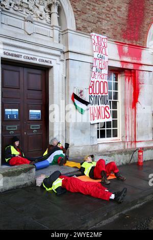 Palästinensische aktionäre sitzen in Zweiergruppen bei einer Demonstration vor dem Rathaus des Bezirks Somersetís. Unterstützer der Palästinensischen Aktion blockieren den Eingang zur County Hall mit Sperren und sprühen rote Farbe auf das Äußere des Gebäudes. Die Demonstranten fordern, dass der Stadtrat von Somerset den Mietvertrag für Büroflächen beendet, die sie an Elbit Systems in Aztec West, Bristol, vermieteten. Sie argumentieren, dass die von Elbit Systems in Großbritannien hergestellten Waffen von der israelischen Verteidigungsstreitkräfte gegen Palästinenser in Gaza und anderswo eingesetzt werden. Bei den israelischen Bombenanschlägen im Gazastreifen sind seit Oktober 2023 mehr als 30.000 Palästinenser ums Leben gekommen. Blass Stockfoto