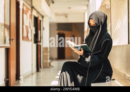 Die junge muslimische Niqab-Frau liest den Qur´an und glaubt dem Heiligen Al Qur´an im Universitätsgebäude. Arabische schwarze Chador-Dame. Stockfoto