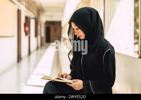 Die junge muslimische Niqab-Frau liest den Qur´an und glaubt dem Heiligen Al Qur´an im Universitätsgebäude. Arabische schwarze Chador-Dame. Stockfoto