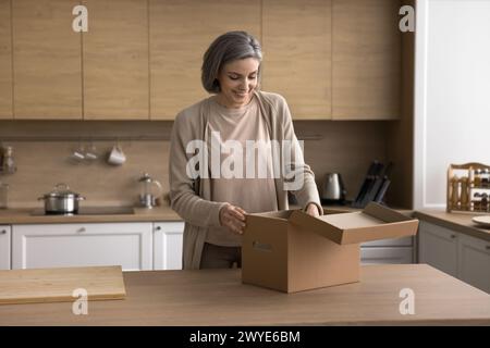 Positive reife Frau, die am Küchentisch steht und das Paket auspackt Stockfoto