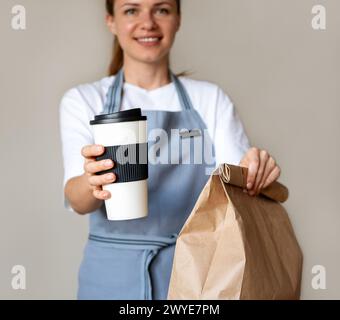 Kompostierbare Kaffeetasse und Papiertüte mit Essen zum Mitnehmen in den Händen einer hübschen Kellnerin. Umweltfreundliches Paket für Speisen zum Mitnehmen. Stockfoto