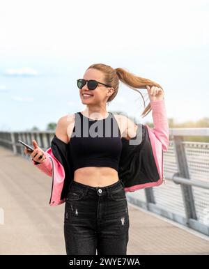 Unbeschwert inspirierte, urbane Frau mit Sonnenbrille und rosa Lederjacke, die einen Spaziergang in der Stadt macht. Stockfoto