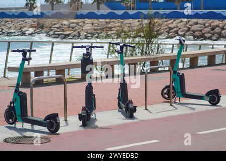 Drei Elektroroller parkten entlang des Geländes einer Promenade, mit dem Meer und den Felsen im Hintergrund, bereit, gemietet zu werden und die Küste zu erkunden Stockfoto