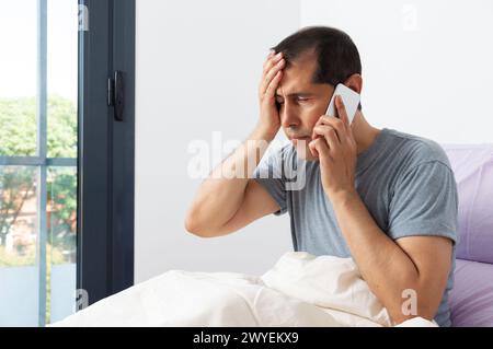 Foto von kranken und Kopfschmerzen Geschäftsmann auf dem Bett sitzen und Arzt im Schlafzimmer anrufen Stockfoto