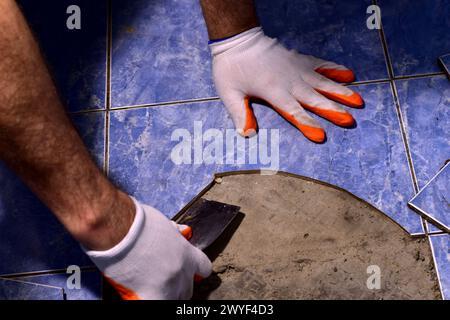 Auf dem Bild sind Männerhände in Handschuhen mit einem Spachtel an der Reparatur des Keramikbodens beteiligt. Stockfoto