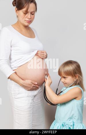 Neugieriges Mädchen, das Stethoskop am Bauch ihrer schwangeren Mutter benutzt, während sie vor neutralem Hintergrund zusammenstehen. Moment der Familienbindung und an Stockfoto