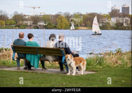 Hamburg, Deutschland. April 2024. Die Menschen auf einer Parkbank genießen die angenehmen Temperaturen auf der Außenalster, während ein Segelboot auf dem Wasser vorbeifährt. Quelle: Georg Wendt/dpa/Alamy Live News Stockfoto