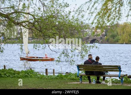 Hamburg, Deutschland. April 2024. Die Menschen auf einer Parkbank genießen die angenehmen Temperaturen auf der Außenalster, während ein Segelboot auf dem Wasser vorbeifährt. Quelle: Georg Wendt/dpa/Alamy Live News Stockfoto