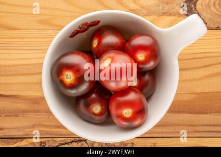 Mehrere saftige schwarze Kirschtomaten in einer weißen Keramikschale auf einem Holztisch, Makro, Blick von oben. Stockfoto