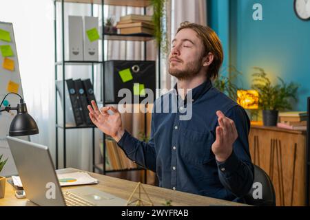 Friedlich entspannter junger männlicher Geschäftsmann, der am Arbeitsplatz mit gefalteten Mudra-Händen sitzt, Stress während des Arbeitstages reduziert, meditiert oder Yoga-Atemübungen im Home Office macht Stockfoto
