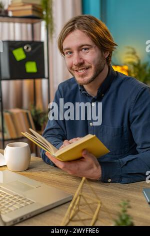 Junger Geschäftsmann, der interessante Bücher liest, lächelnd Seiten umblättert und Literatur genießt, sich nach der Arbeit ausruht. Glücklicher fröhlicher Freelancer mit Laptop, der sich am Tisch zu Hause im Büro entspannt. Vertikal Stockfoto