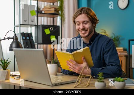 Kaukasischer junger Geschäftsmann liest interessantes Buch, dreht lächelnd Seiten und genießt Literatur, ruht sich nach der Arbeit aus. Glücklicher fröhlicher Freelancer mit Laptop, der sich am Tisch zu Hause im Büro entspannt. Stockfoto