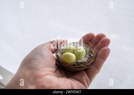 Detail einer Hand, die ein kleines Vogelnest mit drei kleinen grünen Ostereiern hält, gefüllt mit Schokolade. Es ist ein Osterbonbon-Tisch mit dem typischen Stockfoto