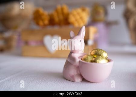 Detail eines kleinen Porzellan-Osterhasen mit einem Korb mit Eiern auf einem Osterbonbon-Tisch mit Ostereiern, Schokolade, Kuchen, Süßigkeiten und Häschen. Stockfoto