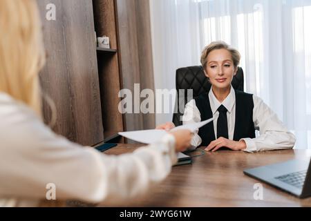 Porträt einer lächelnden Geschäftsmanagerin mittleren Alters, die Papiere an eine nicht erkennbare Kollegin weitergibt, die am Tisch im Büro sitzt. Stockfoto
