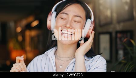 Glückliche asiatische Frau, Kopfhörer und Playlist-Musik im Café zum Audio-Streaming, Tanzen oder Singen. Weibliche Person oder Freiberufler lächeln oder Stockfoto