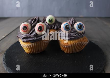 Cupcakes Kuchen mit schwarzgrauer Creme und Augen auf grauem Hintergrund mit Platz für Text. Vorbereitung des Konzepts des Halloween-Feiertags. Stockfoto