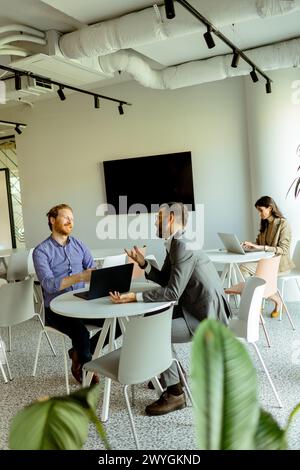 Zwei Männer führen ein lebhaftes Gespräch an einem Tisch, während eine Frau im Hintergrund unabhängig arbeitet Stockfoto