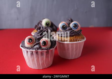 Drei Cupcakes mit Augen auf rotem Hintergrund. Lustige Kuchen Für Kinder Halloween-Party Mit Platz Für Copy Space Text. Stockfoto