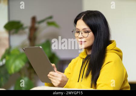 Eine Frau mit Brille schaut sich ein Tablet an. Sie trägt einen gelben Hoodie. Das Tablet ist ein Apple-Produkt Stockfoto