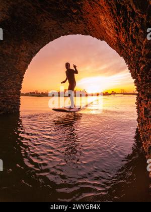 Tragflächenreiter gleitet mit seinem Brett in einem der Kanäle der Ria de Aveiro in Portugal während des Sonnenuntergangs über dem Wasser. Stockfoto