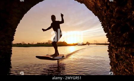 Tragflächenreiter gleitet mit seinem Brett in einem der Kanäle der Ria de Aveiro in Portugal während des Sonnenuntergangs über dem Wasser. Stockfoto