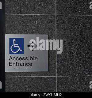 Rechteckiges, barrierefreies Eingangsschild mit blauem Rollstuhlsymbol, schwarzem Text, brailleschrift und schwarzem Pfeil an einer dunklen Wand Stockfoto
