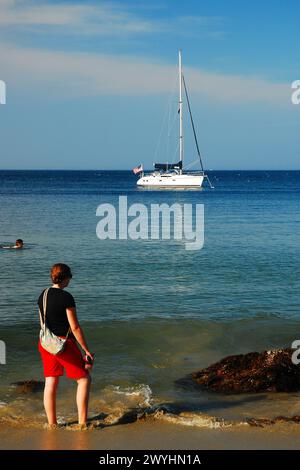 Eine junge Frau schaut auf ein Segelboot, das an einem Sommertag vor der Küste des Strandes liegt Stockfoto