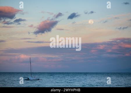 Ein Segelboot liegt irgendwo vor dem Strand von Kihei, während das Sonnenaufgangslicht die Wolken rosa färbt und der Vollmond am Morgenhimmel über Maui sitzt Stockfoto