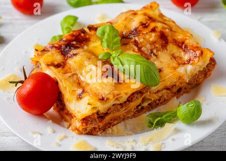 Hausgemachte Fleischlasagne mit frischem Basilikum, Parmesankäse und Kirschtomaten auf einem Teller auf weißem Holztisch. Nahaufnahme. Leckeres italienisches Essen für Stockfoto