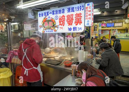 Nachtmarktszene mit Köchen, die kochen, und Gästen, die an einem belebten Imbissstand mit bunten Schildern sitzen Stockfoto