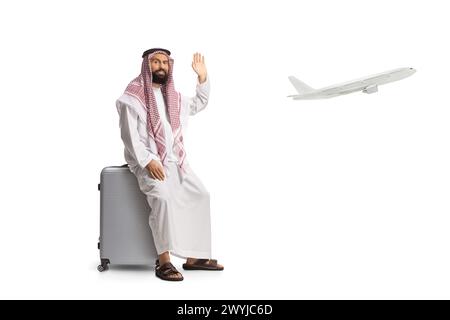 Saudi-arabischer Mann sitzt auf einem Koffer am Flughafen und winkt isoliert auf weißem Hintergrund, Flugzeug fliegt hinten Stockfoto